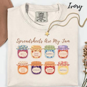 K&ouml;nnte beinhalten: Elfenbeinfarbenes T-Shirt mit dem Aufdruck "Spreadsheets Are My Jam" und bunten Marmeladenglas-Grafiken, die Tabellenkalkulationsfunktionen wie IF() und VLOOKUP() darstellen. Eine goldene Stern-Anh&auml;nger-Halskette liegt auf dem Shirt.