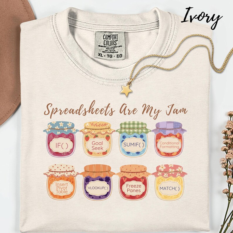 K&ouml;nnte beinhalten: Elfenbeinfarbenes T-Shirt mit dem Aufdruck "Spreadsheets Are My Jam" und bunten Marmeladenglas-Grafiken, die Tabellenkalkulationsfunktionen wie IF() und VLOOKUP() darstellen. Eine goldene Stern-Anh&auml;nger-Halskette liegt auf dem Shirt.