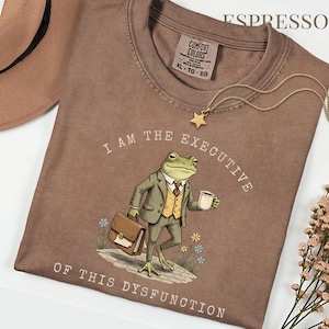 Puede incluir: Camiseta color espresso con el texto "I AM THE EXECUTIVE OF THIS DYSFUNCTION." El gr&aacute;fico presenta una rana con traje sosteniendo un malet&iacute;n y una taza de caf&eacute;. Un collar de estrella dorada est&aacute; sobre la camiseta.