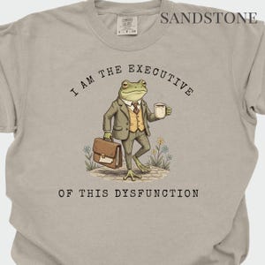 Puede incluir: Camiseta color arena con el texto "I AM THE EXECUTIVE OF THIS DYSFUNCTION". El gr&aacute;fico presenta una rana con traje sosteniendo un malet&iacute;n y una taza de caf&eacute;. La camiseta tiene una etiqueta Comfort Colors.