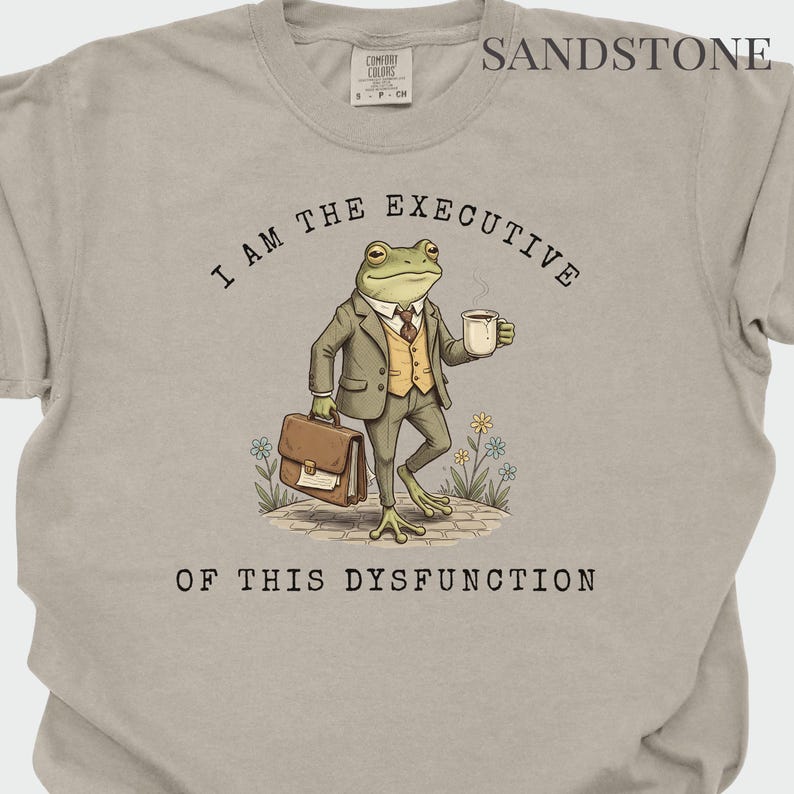 Puede incluir: Camiseta color arena con el texto "I AM THE EXECUTIVE OF THIS DYSFUNCTION". El gr&aacute;fico presenta una rana con traje sosteniendo un malet&iacute;n y una taza de caf&eacute;. La camiseta tiene una etiqueta Comfort Colors.