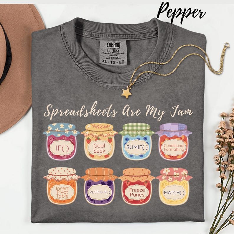 K&ouml;nnte beinhalten: Ein graues T-Shirt mit dem Text "Spreadsheets Are My Jam" und bunten Marmeladenglas-Grafiken, die Tabellenkalkulationsfunktionen darstellen. Eine goldene Sternanh&auml;nger-Halskette ist &uuml;ber dem Shirt drapiert. Das Shirt ist mit XL-TG-EG gekennzeichnet.