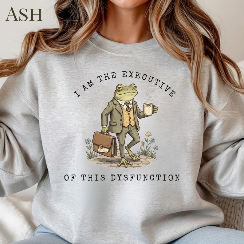 Puede incluir: Sudadera gris claro con el texto "I AM THE EXECUTIVE OF THIS DYSFUNCTION." El gr&aacute;fico presenta una rana de dibujos animados con traje, sosteniendo un malet&iacute;n y una taza de caf&eacute;. La sudadera tiene mangas largas.