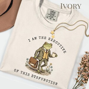 Puede incluir: Camiseta de color marfil con el texto "I AM THE EXECUTIVE OF THIS DYSFUNCTION". El dise&ntilde;o presenta una rana con traje, sosteniendo un malet&iacute;n y una taza de caf&eacute;. Un collar dorado con estrellas est&aacute; sobre la camiseta.