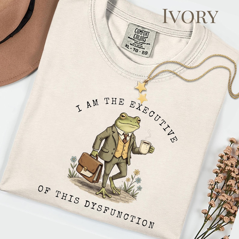 Puede incluir: Camiseta de color marfil con el texto "I AM THE EXECUTIVE OF THIS DYSFUNCTION". El dise&ntilde;o presenta una rana con traje, sosteniendo un malet&iacute;n y una taza de caf&eacute;. Un collar dorado con estrellas est&aacute; sobre la camiseta.