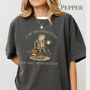 Puede incluir: Camiseta gris oscuro con un dibujo de una rana de dibujos animados con traje, sosteniendo una taza de caf&eacute; y un malet&iacute;n. El texto dice "I AM THE EXECUTIVE OF THIS DYSFUNCTION". La camiseta tiene un corte holgado y una camiseta interior blanca.