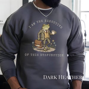 Puede incluir: Sudadera gris oscuro con un dise&ntilde;o humor&iacute;stico de una rana con traje, sosteniendo un malet&iacute;n y una taza de caf&eacute;. El texto dice "I AM THE EXECUTIVE OF THIS DYSFUNCTION". Las palabras "DARK HEATHER" est&aacute;n impresas en la parte inferior.