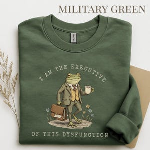 Puede incluir: Sudadera verde militar con el texto "I AM THE EXECUTIVE OF THIS DYSFUNCTION". El gr&aacute;fico presenta una rana con traje sosteniendo una taza de caf&eacute; y un malet&iacute;n. La sudadera es de cuello redondo.