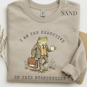 May include: Sand-coloured sweatshirt with the text "I AM THE EXECUTIVE OF THIS DYSFUNCTION." The graphic features a cartoon frog in a suit holding a briefcase and a coffee cup. The frog is standing on a brick path with flowers.