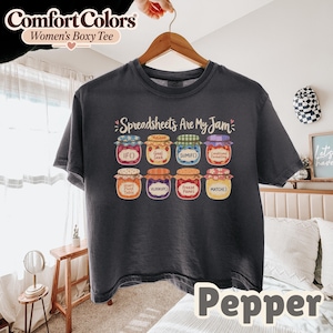 以下が含まれることがあります： チャコールグレーの半袖Tシャツ。8つのカラフルなジャム瓶のグラフィックがプリントされています。各瓶には、IF、Goal Seek、SUMIF、Conditional Formatting、Insert Pivot Table、VLOOKUP、Freeze Panes、MATCHといったスプレッドシートの関数がラベル付けされています。