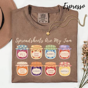 K&ouml;nnte beinhalten: Espressofarbenes T-Shirt mit dem Schriftzug "Spreadsheets Are My Jam" und Illustrationen von Marmeladengl&auml;sern, die jeweils mit Tabellenkalkulationsfunktionen beschriftet sind. Eine goldene Sternanh&auml;nger-Halskette liegt auf dem Shirt.