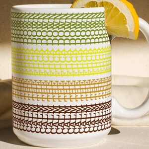 Puede incluir: Una taza de cerámica blanca con un patrón decorativo en tonos verdes, amarillos y marrones. Una rodaja de limón descansa sobre el borde. La taza tiene un asa blanca y está colocada sobre una superficie clara. El diseño de la taza presenta bandas horizontales de patrones repetidos.