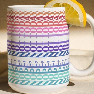 May include: A white ceramic mug with a colorful geometric pattern of repeating shapes and the word "DAY" in a repeating pattern. A lemon slice rests on the rim. The mug is on a light-colored surface.