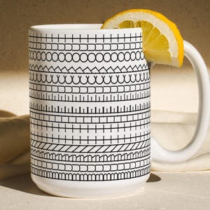 May include: White ceramic mug with a black geometric pattern, a curved handle, and a slice of lemon. The mug's design features various black line patterns. The mug is on a light-colored surface with a folded cloth.