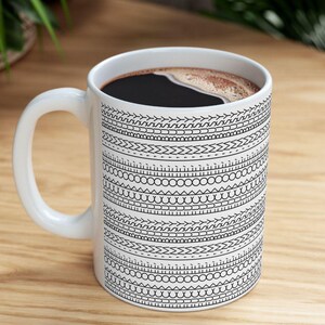 May include: White ceramic coffee mug with a black patterned design filled with dark coffee. The design features horizontal bands of various geometric shapes. The mug sits on a light wooden surface.