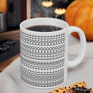 May include: White ceramic mug filled with dark liquid, featuring a repeating black and white geometric pattern. The mug is on a gray surface, with Halloween-themed candies scattered around it. A pumpkin and candles are in the background.