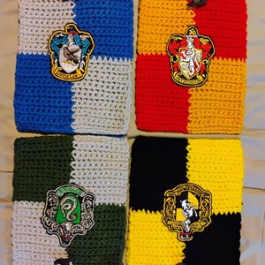May include: Handmade crochet scarves and pouches in various colors, featuring the emblems of four houses. Each scarf and pouch has a unique color scheme, including blue, red, green, and yellow, with matching fringe.