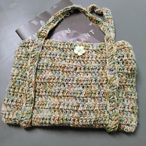 Pode incluir: Bolsa tote de crochê feita à mão em tons de verde, marrom e bege. A bolsa apresenta um botão em forma de flor e alças resistentes. Um livro é parcialmente visível dentro da bolsa. A bolsa é feita de fio texturizado.