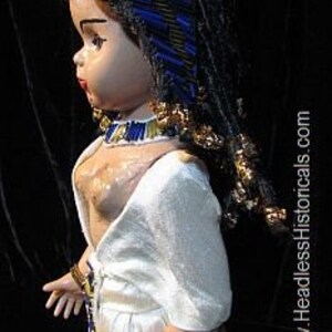 Puede incluir: Una mu&ntilde;eca de porcelana con un tocado azul y dorado, vestida con un vestido blanco con detalles dorados. La mu&ntilde;eca tiene el pelo largo y negro y est&aacute; posada con una mano en la cadera.
