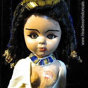 Puede incluir: Una mu&ntilde;eca de porcelana con el pelo negro trenzado, que lleva un vestido blanco y un collar de cuentas azules y doradas. La mu&ntilde;eca tiene los ojos pintados y la boca ensangrentada.