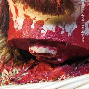 May include: A close-up of a severed head with red paint splattered on it. The head is resting on a woven basket filled with straw. The head is missing its jaw and has a white tooth visible.