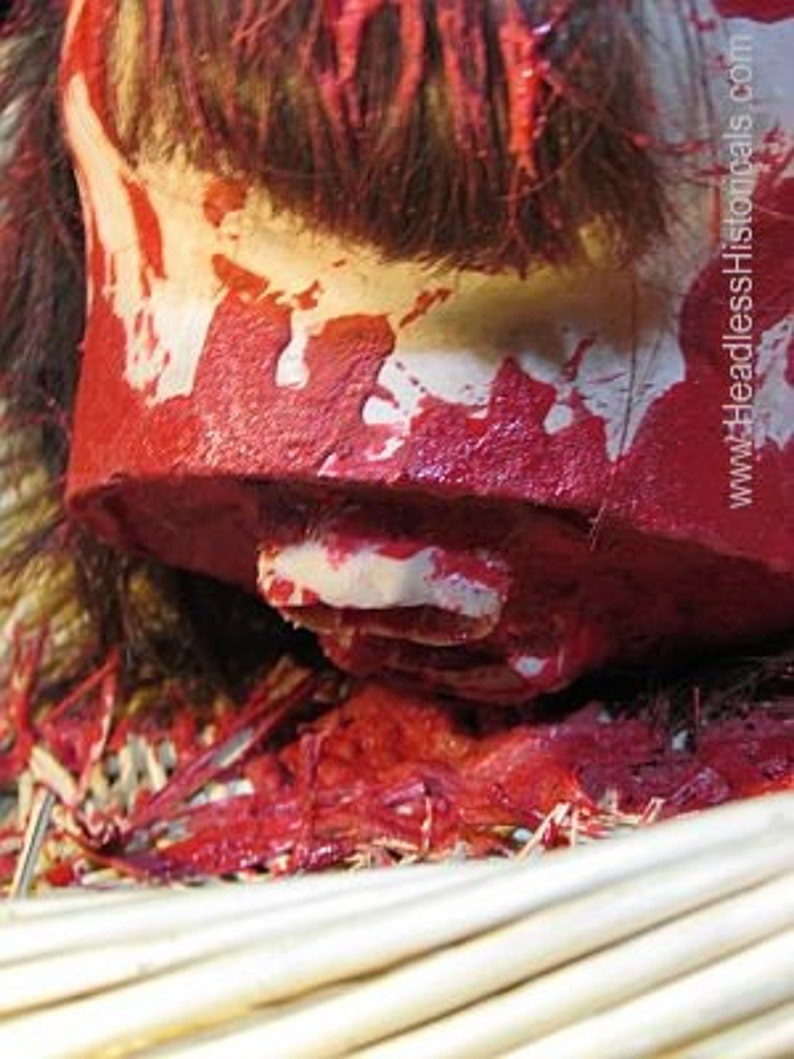 May include: A close-up of a severed head with red paint splattered on it. The head is resting on a woven basket filled with straw. The head is missing its jaw and has a white tooth visible.