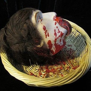 May include: A severed head with long brown hair lies in a wicker basket. The head is covered in blood and the mouth is open, revealing a tongue.
