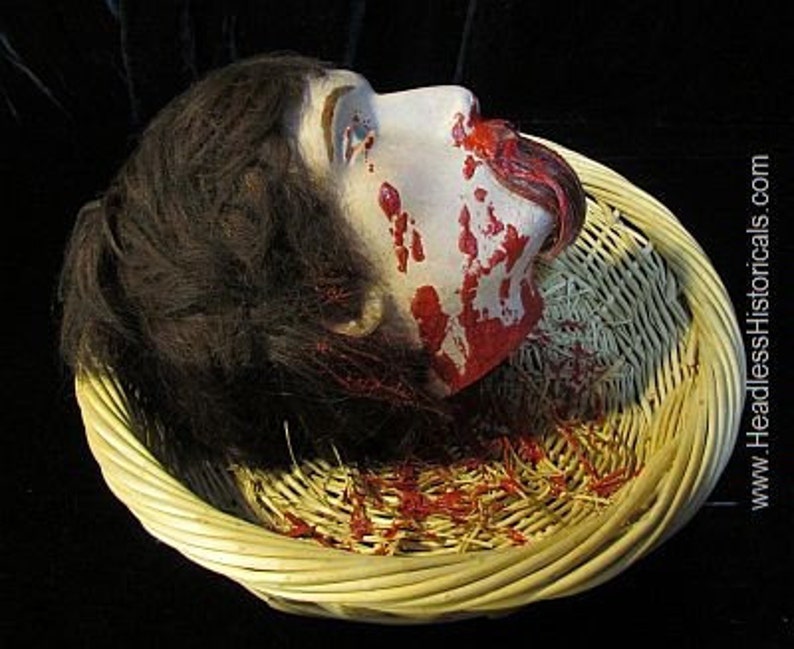 May include: A severed head with long brown hair lies in a wicker basket. The head is covered in blood and the mouth is open, revealing a tongue.