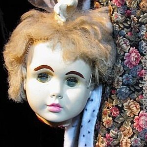 Op de afbeelding: Een vintage porseleinen poppenhoofd met blond haar, blauwe ogen en roze lippen. Het hoofd is los van het lichaam en hangt aan een bloemetjesstof.