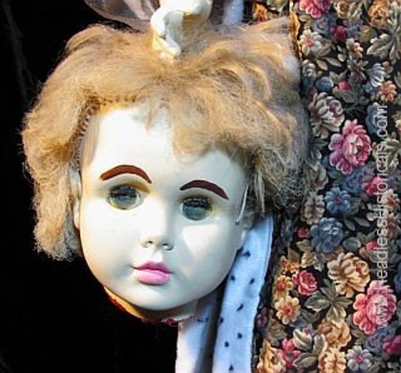 Op de afbeelding: Een vintage porseleinen poppenhoofd met blond haar, blauwe ogen en roze lippen. Het hoofd is los van het lichaam en hangt aan een bloemetjesstof.