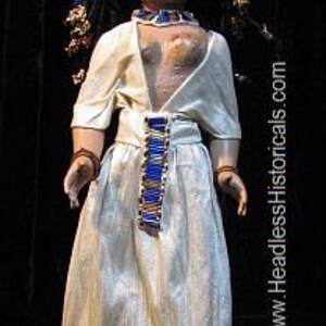 Puede incluir: Una mu&ntilde;eca vestida con un vestido blanco con un cintur&oacute;n azul y dorado. La mu&ntilde;eca tiene el pelo negro y una cara pintada con sombra de ojos azul.