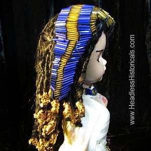Puede incluir: Una mu&ntilde;eca con el pelo largo y casta&ntilde;o que lleva una diadema de cuentas azules y doradas. La mu&ntilde;eca lleva un vestido blanco y un collar de cuentas.