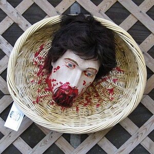 May include: A severed head with fake blood splattered on it is placed inside a woven wicker basket. The head has dark brown hair and a realistic painted face.