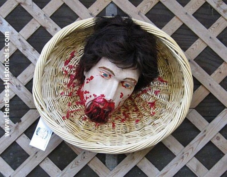 May include: A severed head with fake blood splattered on it is placed inside a woven wicker basket. The head has dark brown hair and a realistic painted face.