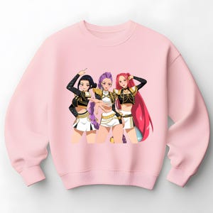 Puede incluir: Una sudadera rosa claro con una colorida ilustración de estilo anime de tres mujeres. Las mujeres visten tops negros y dorados a juego, pantalones cortos blancos y tienen el pelo de diferentes colores: negro, morado y rojo.