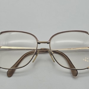 Puede incluir: Un par de gafas de metal en tono dorado con monturas grandes y rectangulares redondeadas. Las varillas son finas y metálicas, y el puente presenta un diseño de doble barra. La varilla interior tiene la inscripción "FRAME JAPAN" en negro.