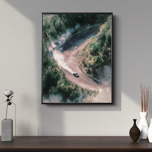 Op de afbeelding: Ingelijste print van een auto die drift op een kronkelende bergweg. De witte auto is omgeven door rook. De weg wordt omzoomd door groene bomen, in een mistige sfeer.
