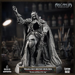 May include: A grey resin miniature of a skeletal warlord figure wearing a crown and tattered cloak, featuring a hollow chest cavity and skull motifs. The base includes a plaque reading WARLORD BRUSH HOLDER and Because painting is the way.