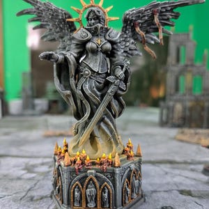 Peut inclure: Une statue miniature détaillée d'une guerrière ange, avec une armure complexe, une épée et de grandes ailes. La statue est grise avec des accents dorés, sur une base décorée avec des bougies. L'ange a un halo et est en pose dynamique.