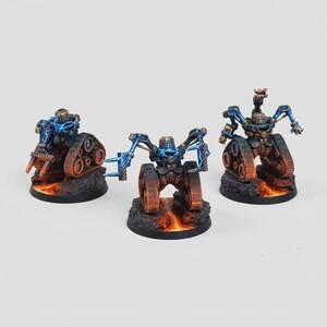 May include: Three miniature robot figures with tank treads and articulated arms. The robots are painted in shades of blue, brown, and gold, with a fiery orange base. The figures are set against a plain white background.