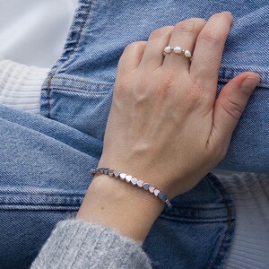 May include: A silver bracelet with heart-shaped links and a ring with three pearls. The jewelry is displayed on a person's wrist and finger, against a backdrop of blue jeans and a gray sweater.