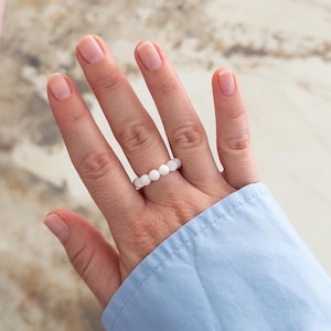 May include: A ring composed of a single row of small, smooth, opaque white spherical beads strung together. The beads are uniform in size and shape, creating a continuous circular band that sits on the finger. The design is simple and monochromatic.