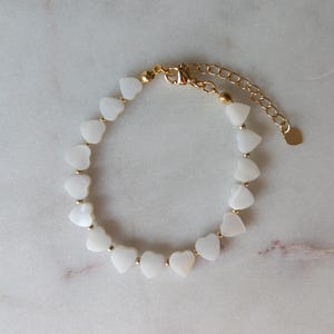 May include: A delicate gold-toned bracelet featuring a series of white heart-shaped beads. The bracelet has a small gold heart charm at the clasp and is set against a marble surface. The bracelet is approximately 7 inches long.