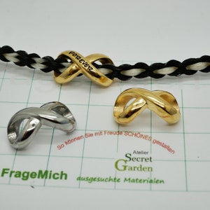 Könnte beinhalten: Zwei Unendlichkeits-Anhänger, einer in Gold und einer in Silber. Der goldene Anhänger hat die Inschrift "Forever" eingraviert. Die Anhänger befinden sich auf einer weißen und schwarzen geflochtenen Schnur.