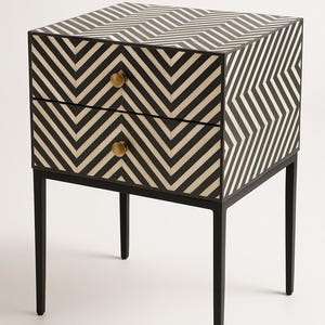 Puede incluir: Una mesita de noche con estampado de chevrón en blanco y negro, con dos cajones y pomos de latón. La mesita de noche tiene una estructura y patas de metal negro. Un mueble elegante para un dormitorio o salón.