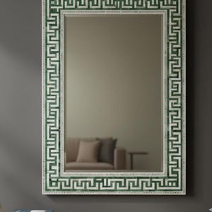 Może przedstawiać: Prostokątne lustro ścienne z biało-zieloną obwódką z motywem greckiego klucza. Lustro odbija pokój z sofą. Rama to połączenie bieli i zieleni, tworzące element dekoracyjny. To lustro to stylowy dodatek do każdego wystroju wnętrz.