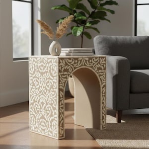 Può includere: Un tavolino beige con un motivo floreale bianco. Il tavolo ha un'apertura ad arco ed è sormontato da un vaso di fiori secchi e libri. Il tavolo è alto 51 cm, largo 46 cm e profondo 46 cm.