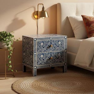 Op de afbeelding: Een marineblauwe nachtkast met twee lades en een wit bloemmotief. De nachtkast heeft messing knoppen en staat naast een bed met een wit hoofdbord. Boven de nachtkast is een goudkleurige wandlamp gemonteerd.