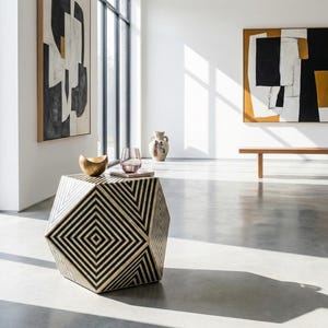 Puede incluir: Mesa auxiliar geométrica con diseño de rayas blancas y negras. La mesa tiene forma hexagonal y está coronada con un cuenco dorado, un vaso y un libro. La habitación presenta arte abstracto y una paleta de colores neutros. La mesa mide unos 45 cm de alto.