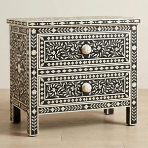 Puede incluir: Una mesita de noche en blanco y negro con dos cajones. Los cajones y los lados presentan un diseño floral y geométrico. Los pomos son blancos y redondos. La mesita es un mueble decorativo.